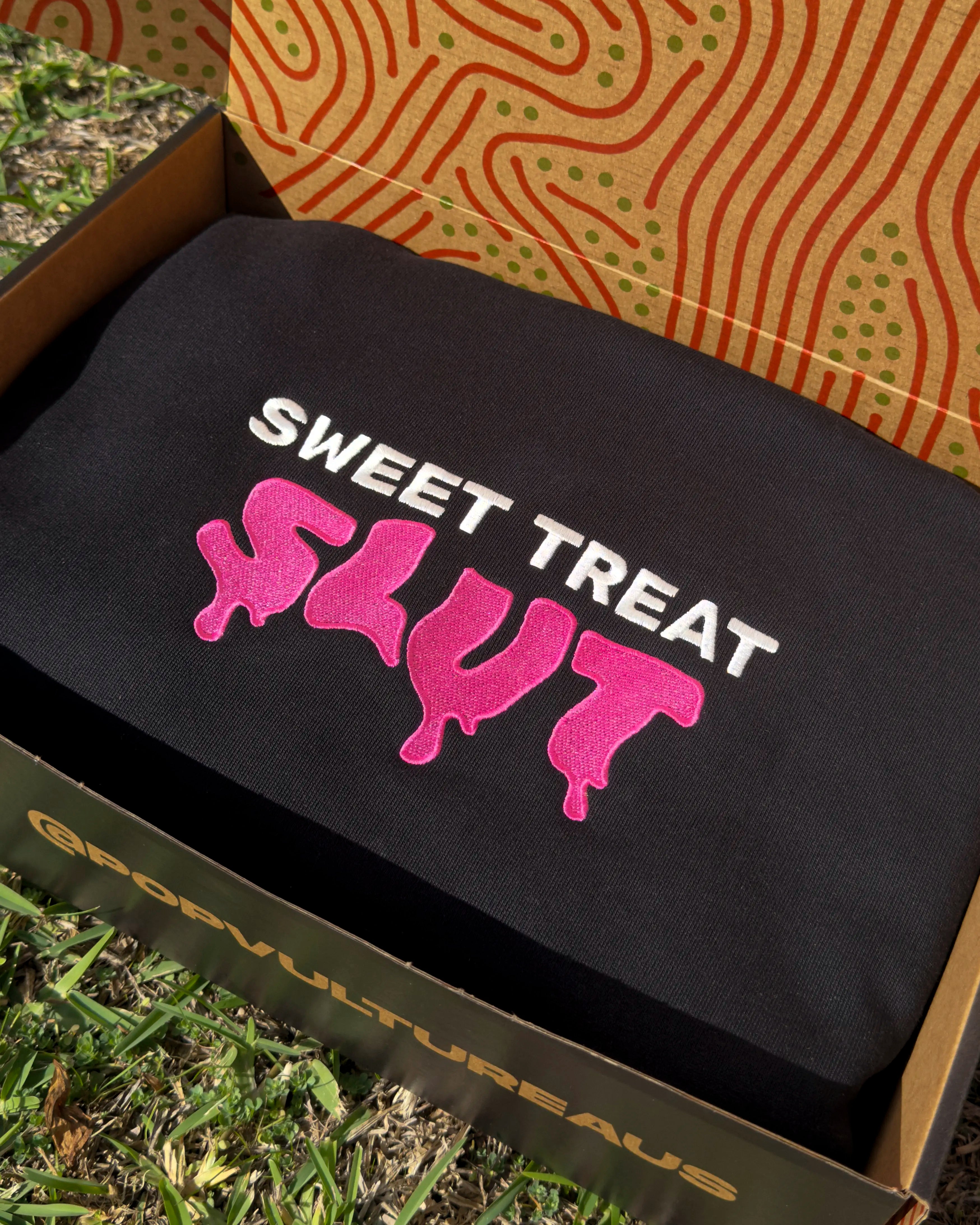 Black hoodie with 'Sweet Treat Slut' text in a yellow box on grass