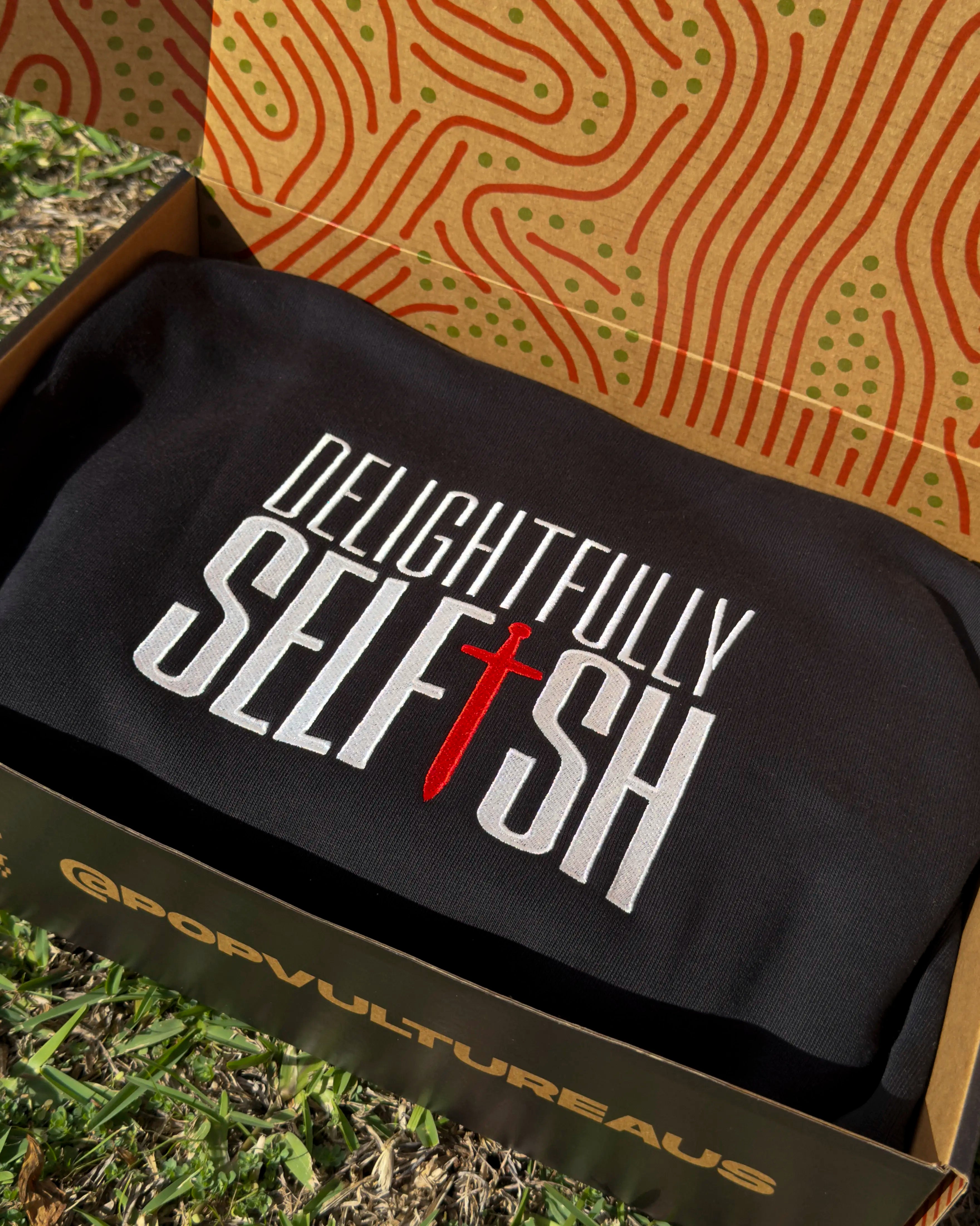 Black hoodie with 'Delightfully Selfish' text in a yellow box on grass