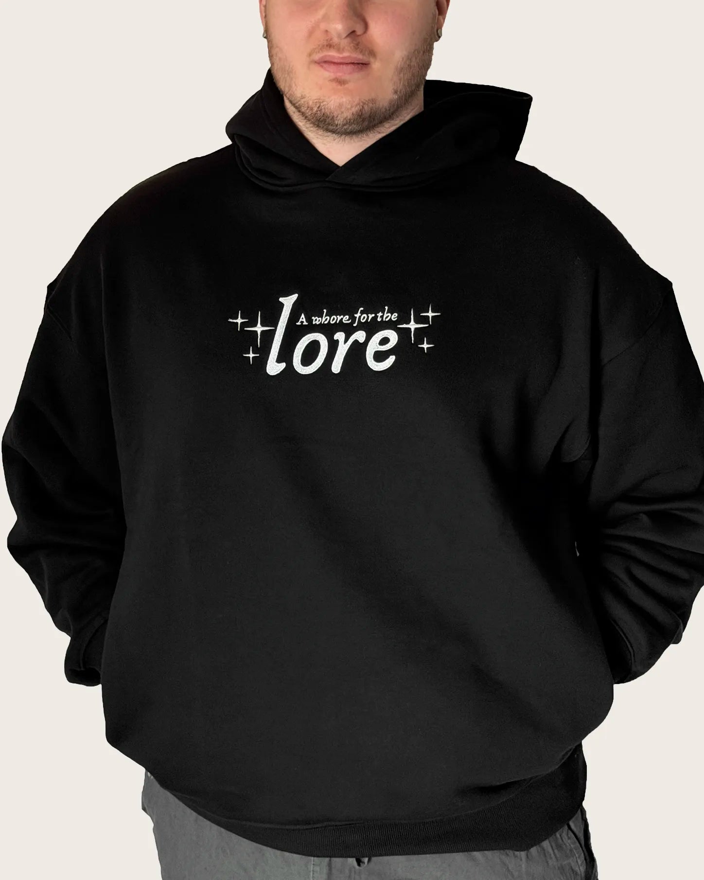 Close up of man wearing oversized black hoodie with embroidered text 'a whore for the lore'.