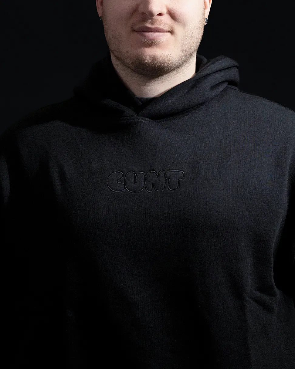 A close-up of a man wearing a black hoodie with embroidered text 'cunt' against a solid black background.