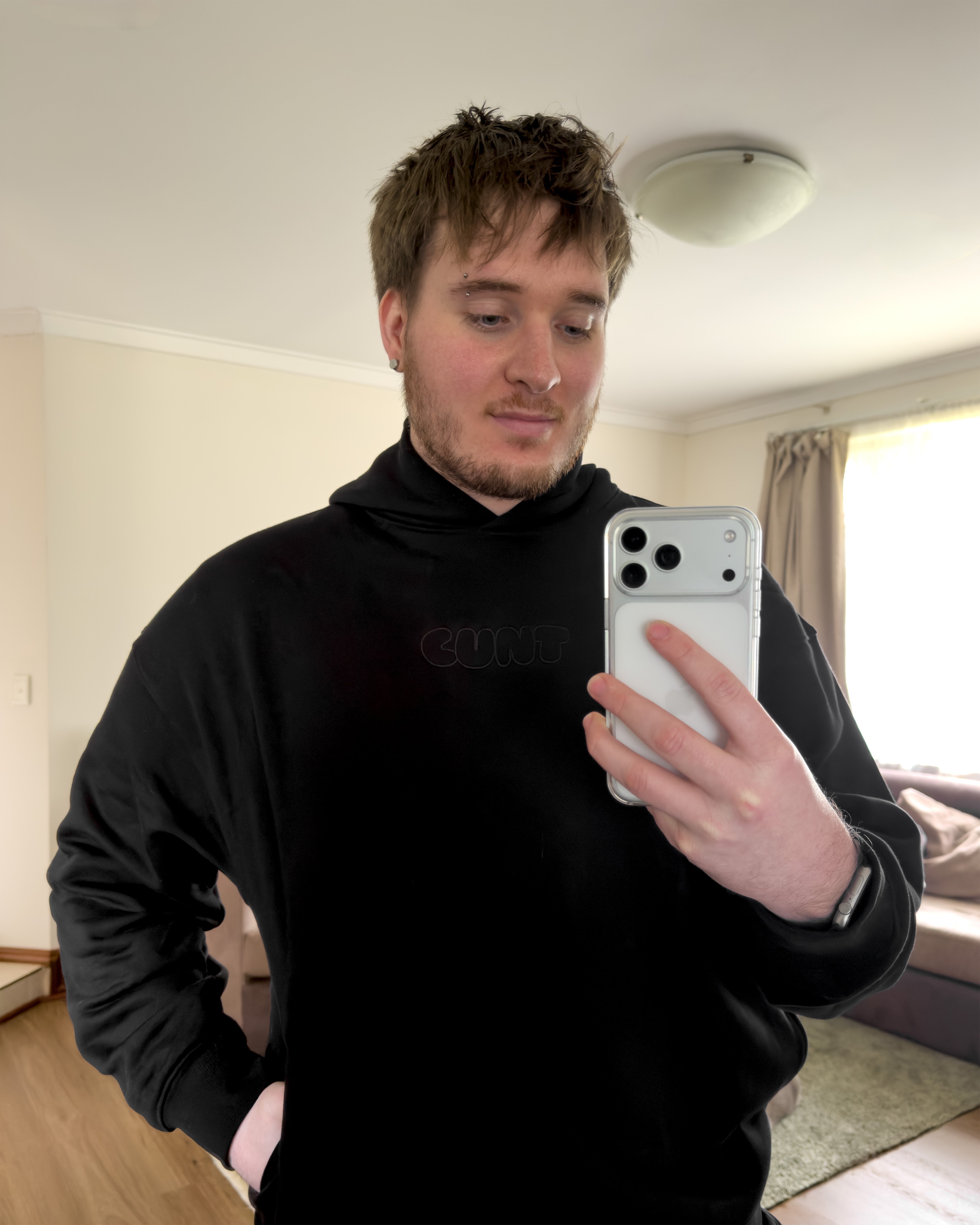 A man taking a mirror selfie in a room, wearing a black hoodie with a subtle cunt logo on the chest and holding a white smartphone.