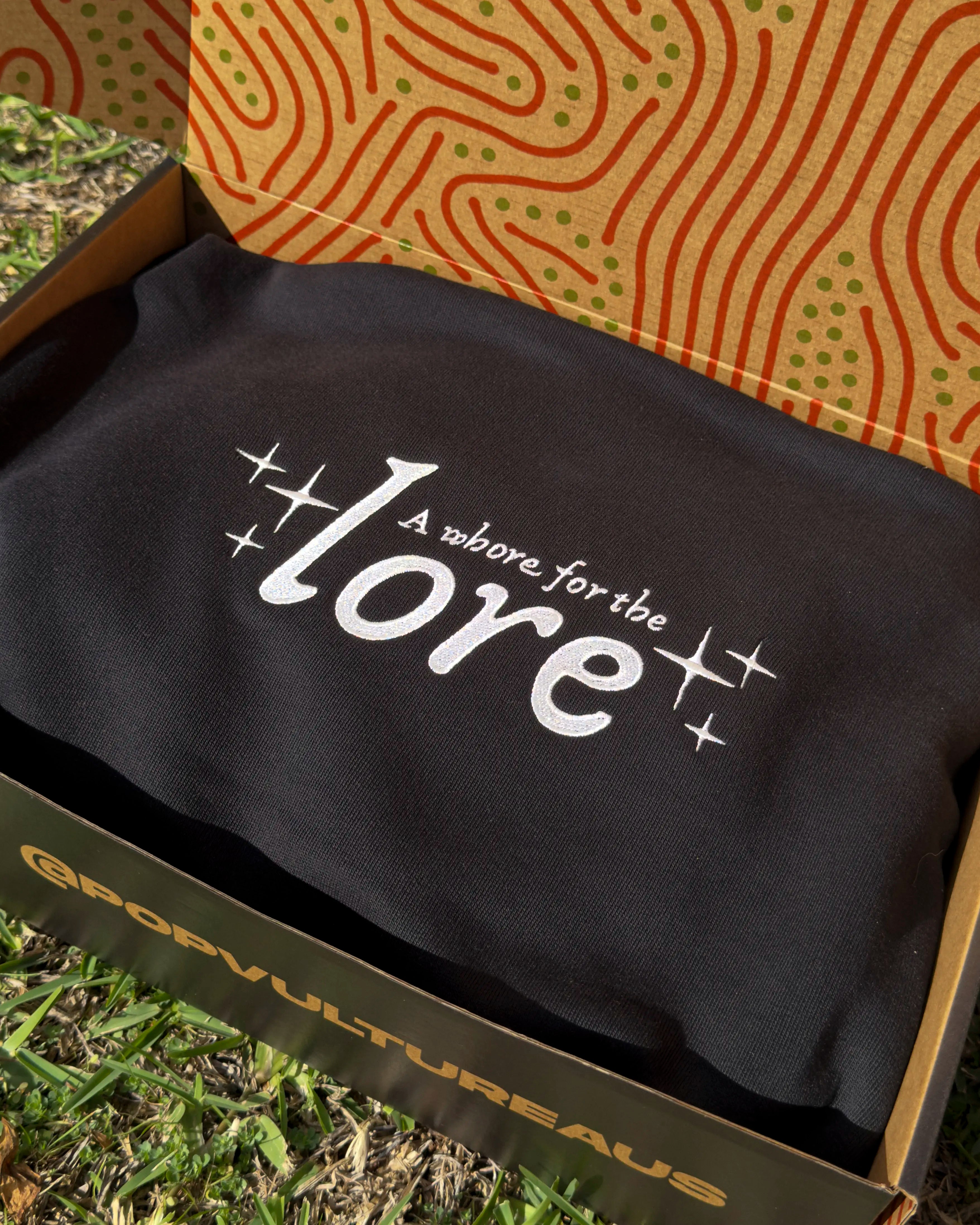 Black hoodie with 'A whore for the lore' text in a yellow box on grass