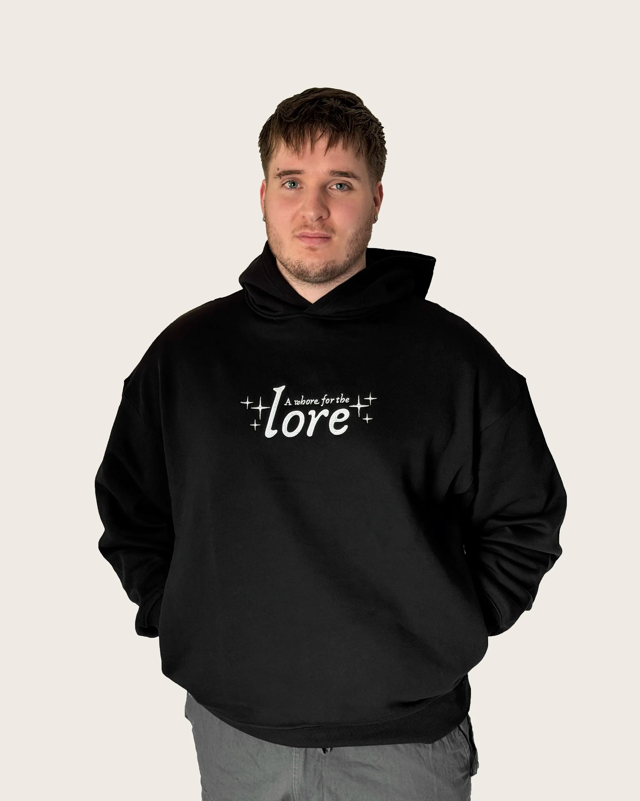Man wearing oversized black hoodie with embroidered text 'a whore for the lore'.