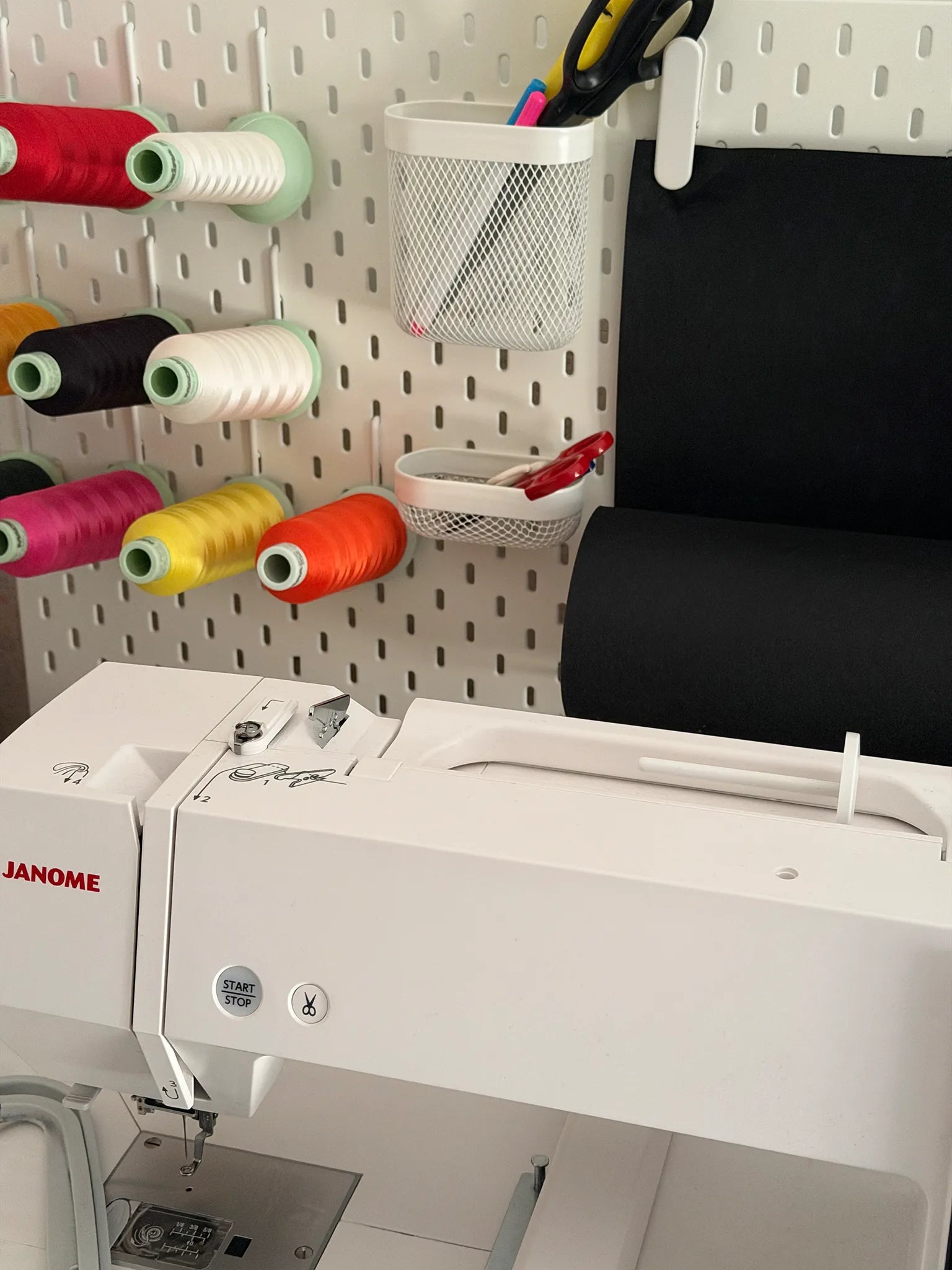 A white Janome embroidery machine in the foreground against a white pegboard wall with colorful thread spools, a mesh pen holder, and a black fabric roll.